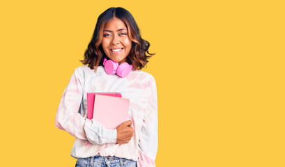 Fototapeta premium Young beautiful mixed race woman holding books wearing headphones looking positive and happy standing and smiling with a confident smile showing teeth