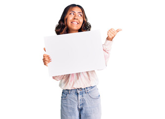 Young beautiful mixed race woman holding cardboard banner with blank space pointing thumb up to the side smiling happy with open mouth