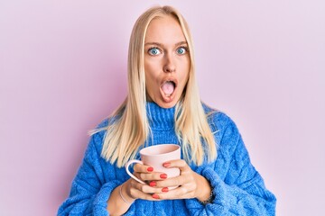 Young blonde girl wearing winter sweater and drinking a cup of hot coffee afraid and shocked with...