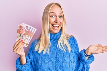 Young blonde girl holding hong kong 10 dollars banknotes celebrating achievement with happy smile and winner expression with raised hand