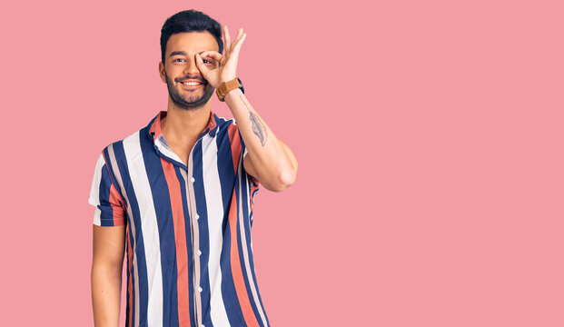 Young handsome hispanic man wearing casual  summer shirt smiling happy doing ok sign with hand on eye looking through fingers
