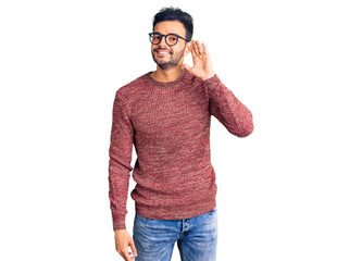 Fototapeta premium Young handsome hispanic man wearing winter sweater and glasses smiling with hand over ear listening an hearing to rumor or gossip. deafness concept.