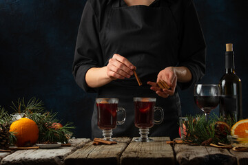 Professional chef adds cinnamon stick to glasses with mulled wine on rustic wooden table with festive composition background. Backstage of cooking hot drink with fragrant spices. Frozen motion.