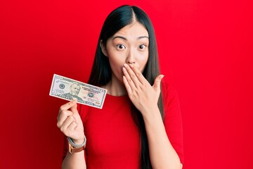 Young chinese woman holding 50 dollars banknote covering mouth with hand, shocked and afraid for mistake. surprised expression