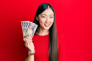 Young chinese woman holding japanese yen banknotes looking positive and happy standing and smiling with a confident smile showing teeth