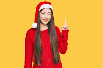 Young chinese woman wearing christmas hat pointing finger up with successful idea. exited and...