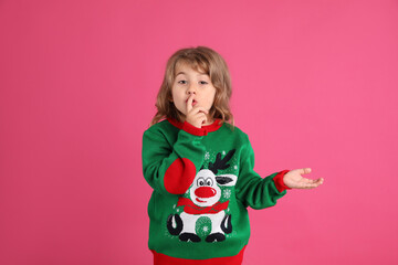 Cute little girl in green Christmas sweater showing silence gesture against pink background