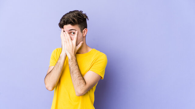 Young caucasian man isolated on purple background blink through fingers frightened and nervous.