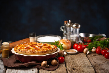 Appetizing meat pie with ingredients for cooking on wooden table on dark blue background. Tasty dinner. Bakery concept. Space for text.