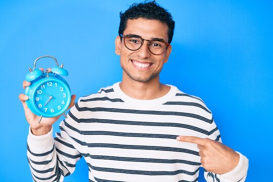 Young handsome hispanic man holding alarm clock pointing finger to one self smiling happy and proud