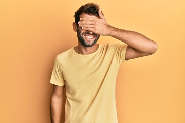 Obraz premium Handsome man with beard wearing casual yellow tshirt over yellow background smiling and laughing with hand on face covering eyes for surprise. blind concept.