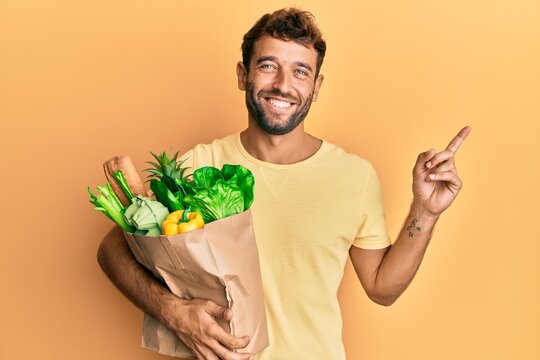 Handsome man with beard holding paper bag with bread and groceries smiling happy pointing with hand and finger to the side