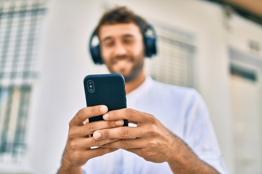 Handsome man with beard wearing casual white shirt on a sunny day smiling happy outdoors using smartphone listening to music wearing headphones