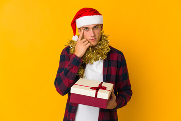 Young caucasian man with christmas hat holding a present isolated on yellow background
