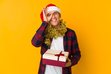 Young caucasian man with christmas hat holding a present isolated on yellow background