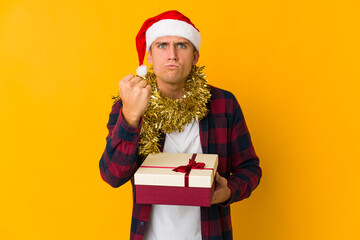 Young caucasian man with christmas hat holding a present isolated on yellow background