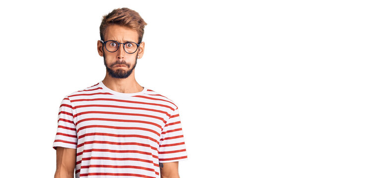 Handsome blond man with beard wearing casual clothes and glasses puffing cheeks with funny face. mouth inflated with air, crazy expression.