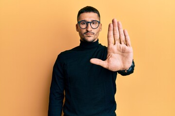 Handsome man with tattoos wearing turtleneck sweater and glasses doing stop sing with palm of the hand. warning expression with negative and serious gesture on the face.