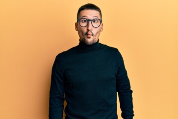 Handsome man with tattoos wearing turtleneck sweater and glasses making fish face with lips, crazy...
