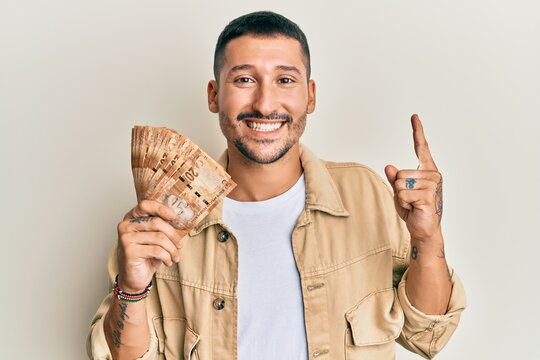 Handsome man with tattoos holding south african 20 rand banknotes smiling with an idea or question pointing finger with happy face, number one
