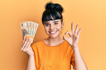 Young hispanic girl holding norwegian krone banknotes doing ok sign with fingers, smiling friendly gesturing excellent symbol