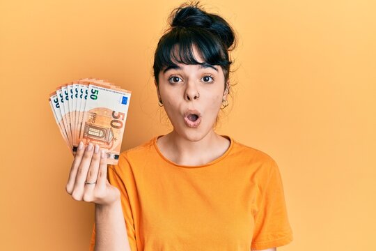 Young hispanic girl holding bunch of 50 euro banknotes scared and amazed with open mouth for surprise, disbelief face