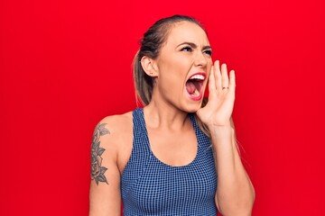 Young beautiful blonde woman wearing sporty t-shirt standing over isolated red background shouting and screaming loud to side with hand on mouth. Communication concept.
