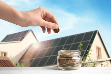 Woman putting coin into jar against house with installed solar panels on roof, closeup. Economic...