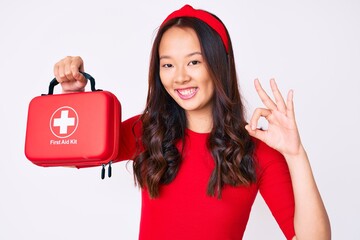 Young beautiful chinese girl holding first aid kit doing ok sign with fingers, smiling friendly...