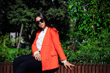 Beautiful brunette woman in red jacket sits on a bench on the street in the park