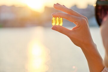 Yellow gel capsule vitamin d omega-3 in womans hand, sun sea background - obrazy, fototapety, plakaty