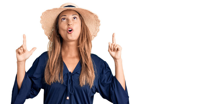 Middle age hispanic woman wearing summer hat amazed and surprised looking up and pointing with fingers and raised arms.