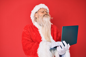 Old senior man with grey hair and long beard wearing santa claus costume using laptop looking at the camera blowing a kiss being lovely and sexy. love expression.