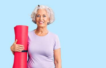 Senior grey-haired woman holding yoga mat looking positive and happy standing and smiling with a confident smile showing teeth