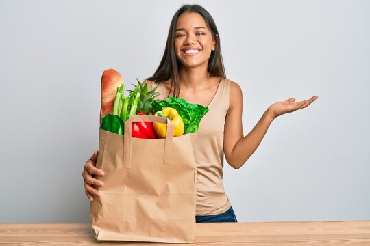 Beautiful Hispanic Woman Holding Paper Bag With Bread And Groceries Celebrating Victory With Happy Smile And Winner Expression With Raised Hands