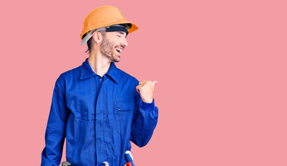 Young hispanic man wearing worker uniform smiling with happy face looking and pointing to the side with thumb up.