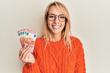 Beautiful blonde woman holding 10 euro banknotes looking positive and happy standing and smiling...