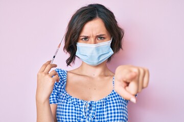 Young beautiful woman wearing medical mask holding syringe pointing with finger to the camera and to you, confident gesture looking serious