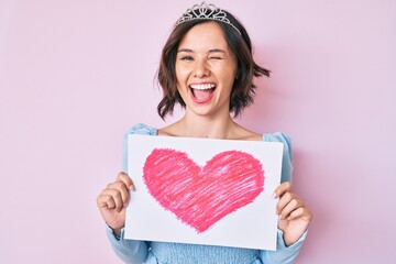 Young beautiful girl wearing princess crown holding heart draw winking looking at the camera with sexy expression, cheerful and happy face.