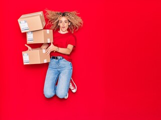 Young beautiful curly deliverywoman holding package smiling happy. Jumping with smile on face over isolated red background.