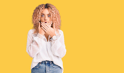 Young blonde woman with curly hair wearing elegant summer shirt shocked covering mouth with hands for mistake. secret concept.