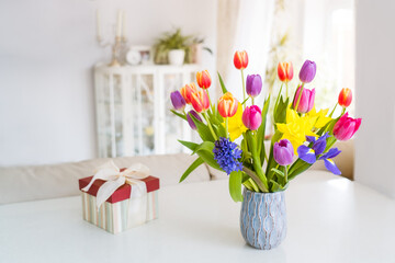 Fresh spring colorful bouquet of tulips, daffodils, irises in vase and gift box on white table with light classic design room background. Festive flowers for gift. Mockup for greeting card. Copy space