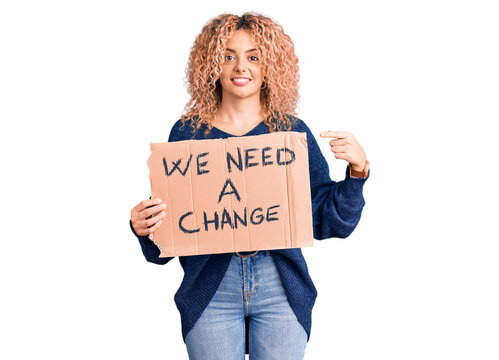 Young blonde woman with curly hair holding we need a change banner smiling happy pointing with hand and finger
