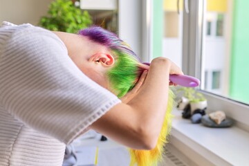 Fashionable teen girl with trendy rainbow dyed hair combing hair at home