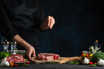 Professional chef pours peppercorns on raw steak on wooden chopped board. Backstage of preparing grilled pork meat at restaurant kitchen on dark blue background. Frozen motion.