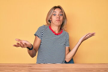 Beautiful caucasian woman wearing casual clothes sitting on the table clueless and confused...