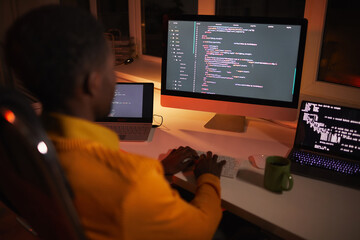 Back view portrait of African-American man writing code while working with multiple computer...