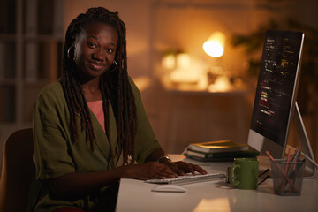 Portrait of contemporary African-American woman writing code and looking at camera while working in...