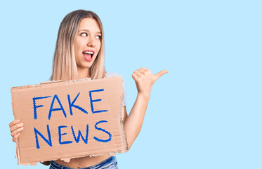 Young beautiful blonde woman holding fake news banner pointing thumb up to the side smiling happy...
