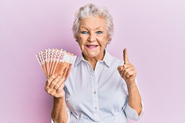 Senior grey-haired woman holding 100 norwegian krone banknotes smiling with an idea or question pointing finger with happy face, number one